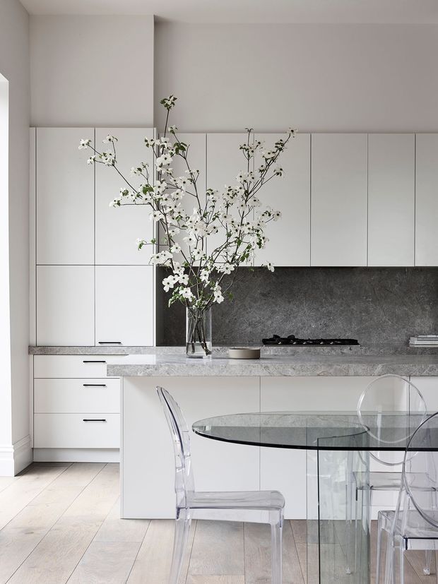 A bright, minimalist kitchen and dining area with full-height white cabinetry, a grey stone benchtop and matching dark grey stone splashback. A curved kitchen island extends into the room, paired with a round glass dining table and transparent acrylic chairs. Pale timber flooring, neutral walls, and delicate flowering branches create a clean, refined atmosphere.