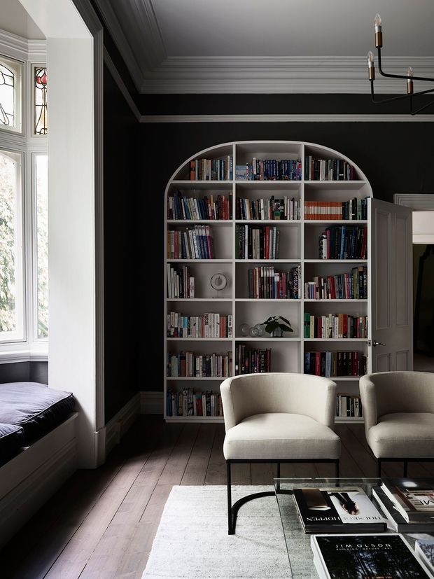 A refined, moody sitting room with a dramatic near-black wall, a large white built-in arched bookcase filled with books, two upholstered armchairs, a glass coffee table, dark timber floorboards and a pale rug. Tall leadlight-style windows and a built-in window seat occupy the left side, while a paneled door opens to another room. Elaborate ceiling cornices and a sculptural dark metal chandelier add architectural and decorative detail.