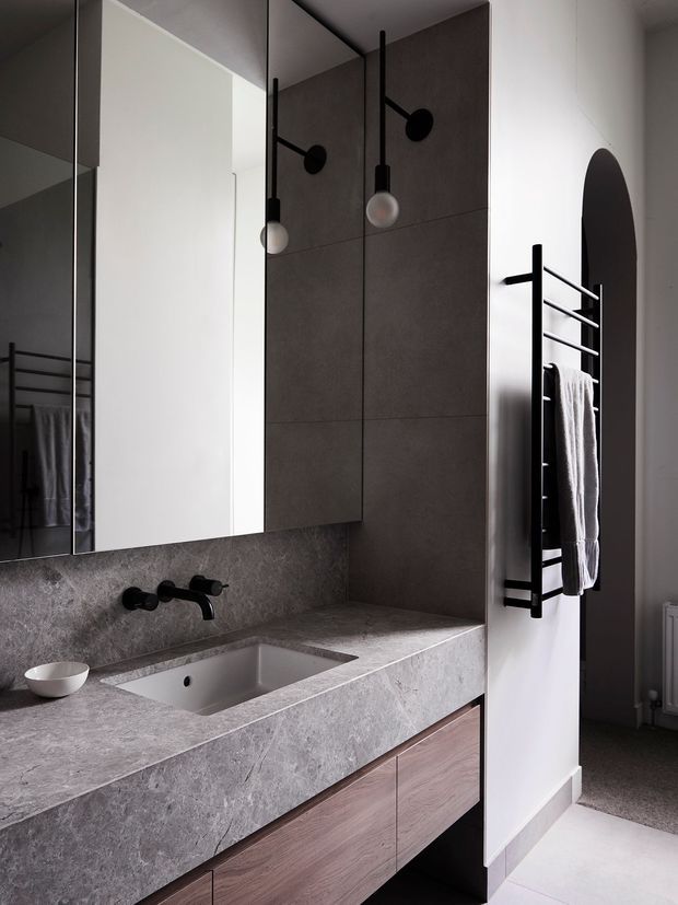 Contemporary bathroom vanity with a wide grey stone benchtop and integrated rectangular basin, dark wall-mounted tapware, large mirrored cabinet, and warm timber drawer fronts. A charcoal tiled feature wall rises behind the vanity, while black pendant lights and a black heated towel rail provide strong graphic accents. The space is refined, minimalist, and softly lit.