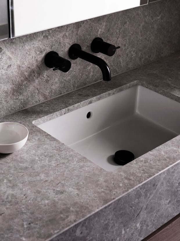 Close-up of a contemporary bathroom vanity featuring a square integrated white basin, matte black wall-mounted tapware, and a grey stone-look benchtop and backsplash. The minimalist fixture arrangement and dark hardware create a refined, monochromatic aesthetic.