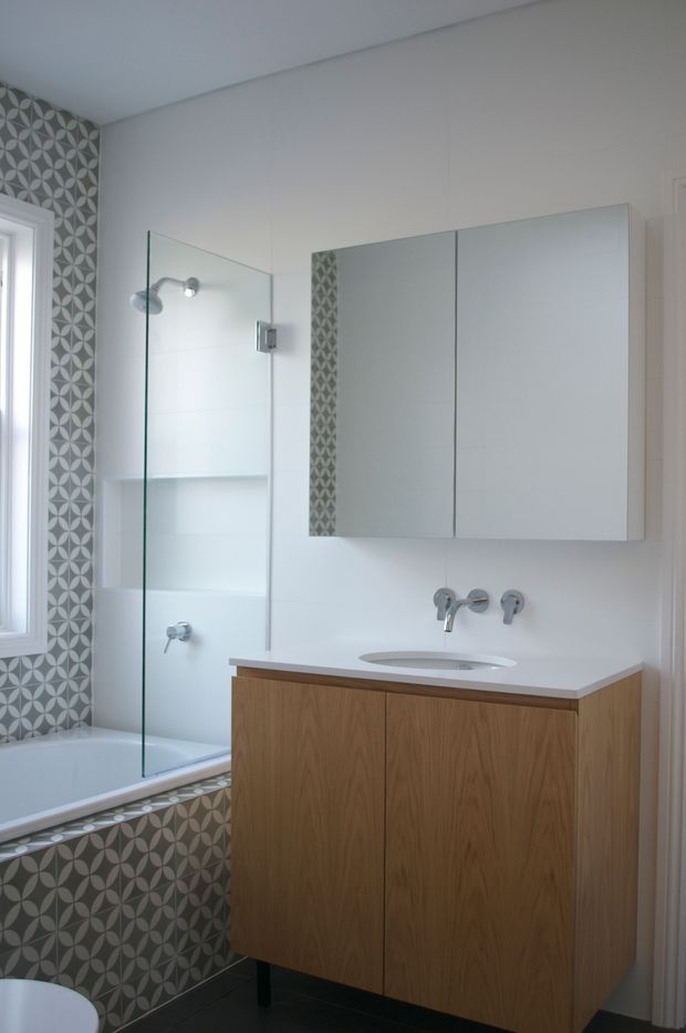 A compact, bright contemporary bathroom with a wall-mounted timber vanity, white integrated basin and chrome wall-mounted taps. A frameless glass screen separates the bathtub and shower area, with recessed white shelving. The walls are mostly white, while a grey and white repeating geometric tile forms a feature wall and bathtub surround. Mirrored wall cabinets and a narrow window add light.