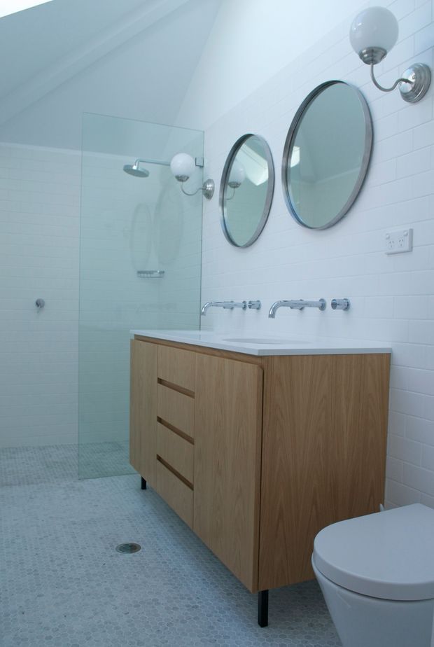 Contemporary bathroom with a wall-mounted timber vanity, dual white basins, chrome wall-mounted taps, round mirrors, white tiled walls, and a frameless glass walk-in shower. Small-format pale mosaic tiles cover the floor, and globe wall sconces provide decorative lighting.