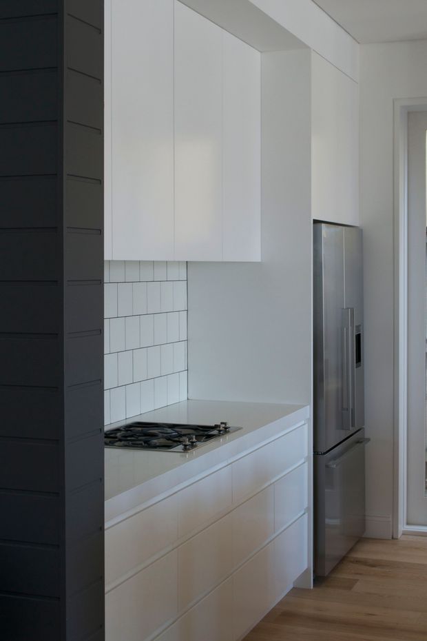 A narrow, contemporary kitchen featuring full-height white handleless cabinetry, white drawers, a pale stone benchtop, and a white rectangular tiled splashback. A black gas cooktop is set into the benchtop, with a tall stainless-steel refrigerator beside the cabinetry. Dark vertical joinery frames the left side, while light timber flooring continues toward a glazed doorway.