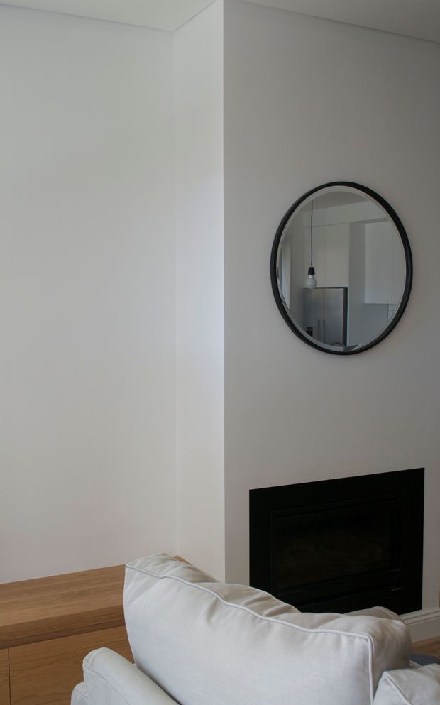 Minimal, softly lit living room corner with white walls, a pale timber floor, a white upholstered sofa in the foreground, and a dark rectangular fireplace integrated into the wall. A round black-framed mirror hangs above the fireplace and reflects a simple pendant light and part of the room.