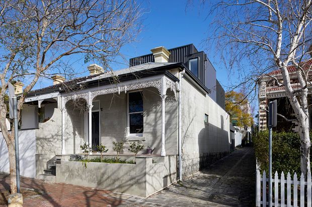 Street-facing view of a narrow urban residence combining a restored historic veranda-fronted facade with a contemporary dark-clad upper-level addition. The facade is light rendered masonry with ornate decorative metal veranda detailing, tall windows, a raised front entry, and a narrow paved passage beside the house. Mature deciduous trees and neighbouring heritage-style homes frame the scene.