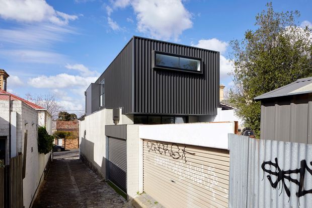 Exterior view of a compact contemporary two-storey dwelling or extension in a dense urban lane. The upper volume is clad in dark vertically ribbed metal with narrow horizontal glazing, while the lower level has white rendered masonry, a roller garage door, and a narrow cobbled access lane. Adjacent sheds, boundary walls, fencing, and mature trees frame the property.