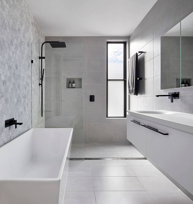 A bright, contemporary bathroom featuring a freestanding white bathtub, walk-in glass shower, wall-mounted white vanity with integrated basin, large mirror, black tapware and hardware, pale grey floor and wall tiles, and a patterned light-grey feature wall. A narrow frosted window provides natural light.
