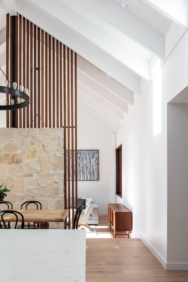 Bright contemporary open-plan interior showing a kitchen island in the foreground, dining area, and living room beyond. A substantial natural stone feature wall is paired with vertical dark timber battens, white pitched and raked ceiling forms, pale timber flooring, and large clerestory-style glazing that fills the space with natural light. The room includes a timber dining table, black pendant lighting, a low timber media console, and a neutral sofa.