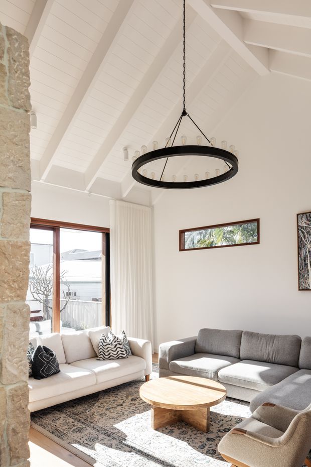 Bright living room with a high vaulted white timber-lined ceiling and exposed beams, a large black circular pendant light, pale walls, natural stone feature wall, timber-framed glazing, and a neutral seating arrangement around a round wooden coffee table. Strong sunlight enters through the windows, creating an airy, relaxed interior.