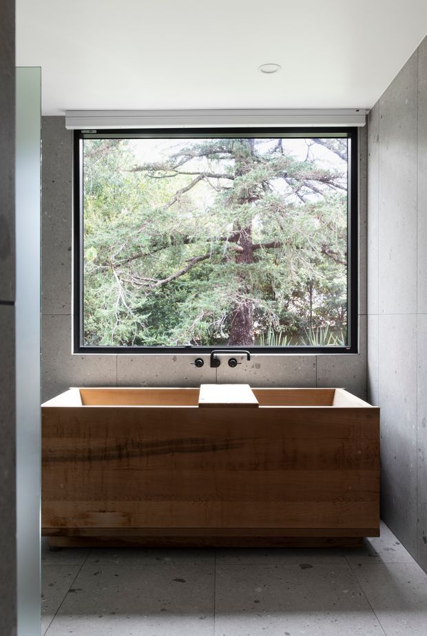 A minimalist bathroom featuring a freestanding timber bathtub positioned beneath a large black-framed window overlooking dense greenery. Grey concrete-look wall and floor finishes create a restrained, contemporary setting, with a wall-mounted bath filler and simple recessed ceiling light.