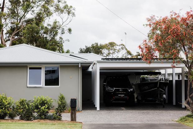 Single-storey suburban dwelling with a light rendered facade, low-pitched corrugated metal roof, and an attached open double carport/garage shelter. The carport contains vehicles, while the front elevation includes simple windows, a paved driveway, and low planted landscaping.