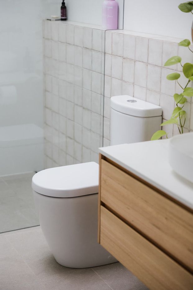 A bright, compact contemporary bathroom featuring a freestanding white bathtub, white toilet, wall-mounted timber vanity with a white benchtop, and a pale square-tiled feature wall. A trailing green plant and toiletries add subtle natural and pastel accents.