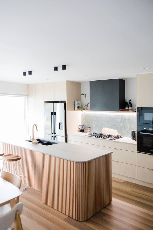 Bright contemporary kitchen featuring a large island with a pale stone benchtop and vertically fluted timber-clad base, light timber cabinetry, integrated stainless-steel appliances, a dark feature cabinet, and a pale small-format tiled splashback. The open-plan space has timber flooring, white walls and ceiling, understated black lighting, and abundant daylight.