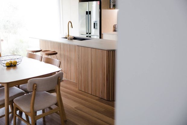 Bright open-plan kitchen and dining area featuring a large island with vertically fluted timber cabinetry, a pale stone benchtop, integrated sink with brass-toned tapware, stainless steel refrigerator, timber flooring, and a light timber dining table with upholstered chairs.
