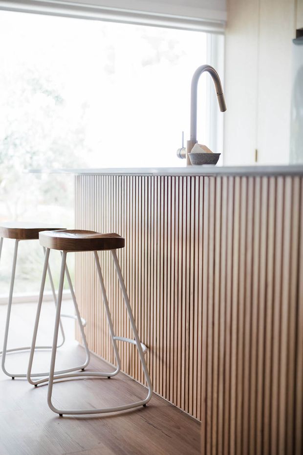 Bright contemporary kitchen island with a vertically slatted timber-clad breakfast bar, pale stone benchtop, tall curved metal tap and two minimalist stools. Large glazing and a light neutral palette create an airy, refined interior.