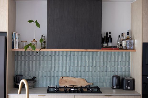 A contemporary kitchen vignette with a pale stone or composite benchtop, black gas cooktop, and a muted blue-green vertically ribbed tile splashback. Light timber cabinetry frames open shelving, which displays books, plants, wine bottles, and spirits. A dark timber feature panel provides contrast above the shelving, while small black appliances and a brushed brass gooseneck tap add functional and decorative details.