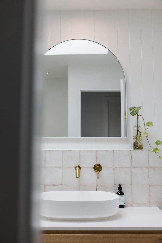 A softly lit contemporary bathroom vanity with a large arched frameless mirror, white vessel basin, wall-mounted brushed brass tapware, pale tiled splashback, and timber-toned vanity cabinetry. A small trailing plant and soap dispenser sit on the counter.