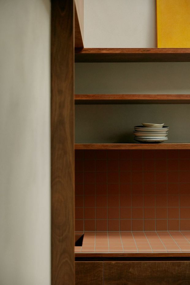 Close-up interior view of a built-in timber shelving and cabinetry unit. Dark walnut-toned vertical framing and shelves surround open storage niches, with a stack of ceramic plates. The lower section features a terracotta-red square tiled backsplash and countertop, with dark timber cabinetry below. The composition is warm, refined, and highly material-focused.