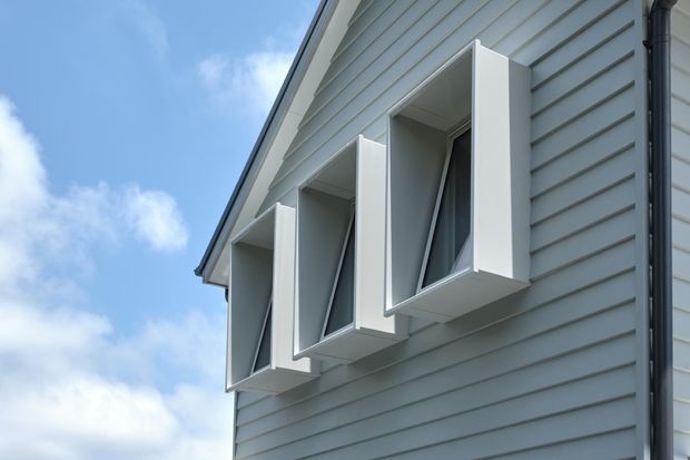 Close-up exterior view of a light grey weatherboard-clad gable wall with three projecting white window surrounds or box bays. Dark-framed windows sit within the deep reveals, and a black vertical downpipe runs along the right edge. The image shows a clean, contemporary treatment of a pitched-roof residential facade against a blue sky.