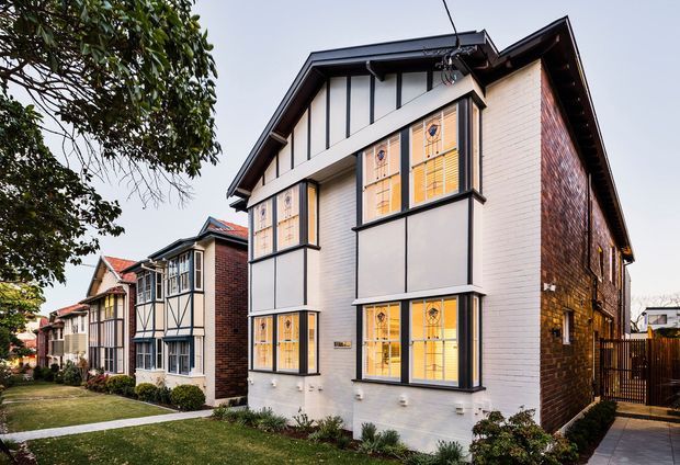 Exterior photograph of a narrow multi-storey attached or semi-attached residence in a suburban streetscape. The foreground house has a steep gabled roof, white painted masonry facade with dark vertical trim and projecting bay windows, patterned leadlight-style glazing, and a red-brown brick side wall. Similar adjoining homes extend along the landscaped frontage, with a paved side path and timber fence visible at right.