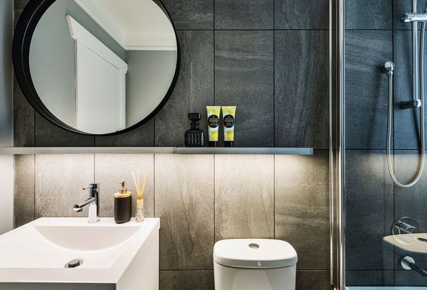 Contemporary bathroom with a wall-mounted white vanity and integrated basin, wall-hung toilet, large round black-framed mirror, and a fully glazed walk-in shower. Dark charcoal stone-look wall panels contrast with a lighter textured backsplash, while recessed shelf lighting adds a refined, atmospheric feel.