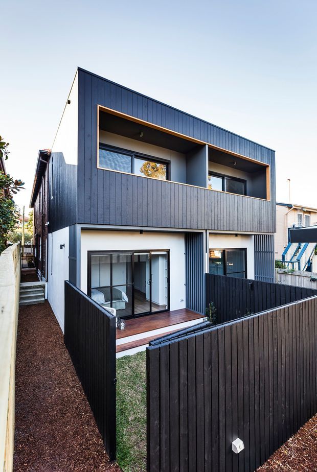 A modern two-storey residential townhouse viewed from the front courtyard. The compact home has dark vertical cladding, a strong geometric box-like form, recessed upper-level balconies, white rendered lower walls, large glazed doors and windows, timber decking, and dark privacy fencing. Narrow landscaped paths and neighbouring homes are visible around the property.