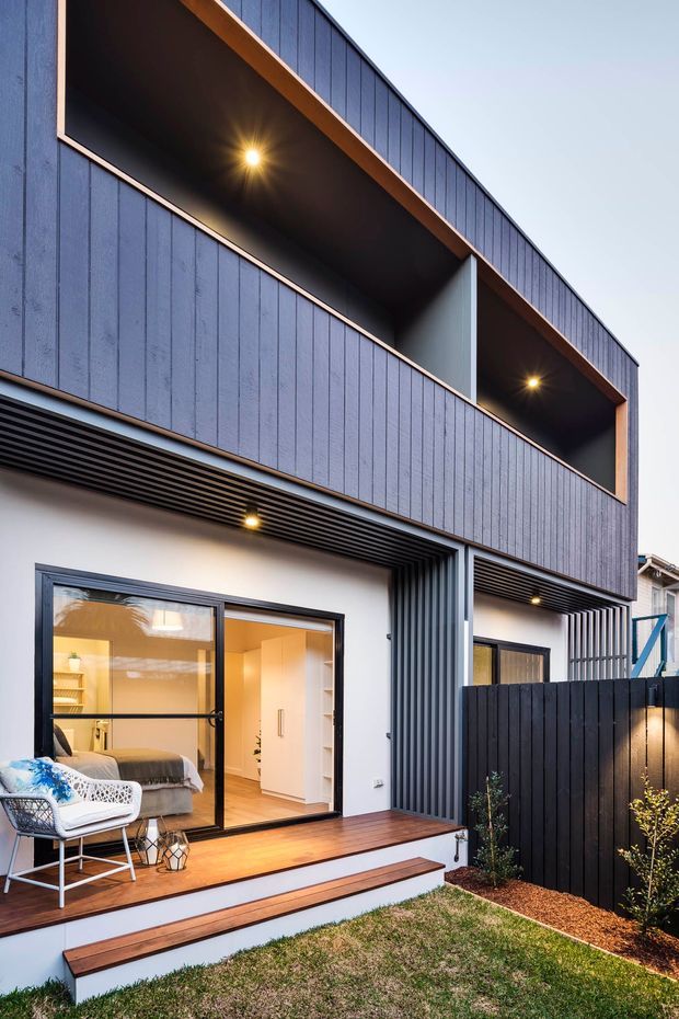 Contemporary two-storey residential facade with dark blue-grey vertical cladding, warm timber trim, and deep recessed upper-level openings with integrated lighting. The lower level opens onto a timber deck and steps through large sliding glass doors, revealing a bedroom and light-toned interior. A dark slatted privacy fence and small landscaped garden occupy the foreground.