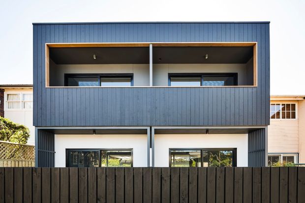 Front exterior view of a contemporary two-storey residential building with a dark vertical-clad upper level, recessed balconies framed in warm timber, large black-framed windows, and an open ground-level frontage partly screened by a dark timber fence. Neighbouring homes are visible on either side.