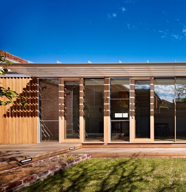 Single-storey residential extension viewed from a landscaped courtyard, featuring a long glazed façade with timber-framed sliding or folding doors, exposed brick walls, a broad horizontal timber pergola or roof overhang, and a timber deck. A timber privacy screen and raised garden edging frame the foreground. The image emphasizes the connection between the interior and outdoor deck, with strong sunlight and a clear blue sky.