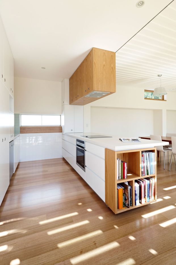 Bright open-plan kitchen and dining area with white flat-panel cabinetry, a large white island, integrated stainless-steel appliances, and a timber overhead rangehood enclosure. The island includes an open timber bookshelf at one end. Warm timber flooring, white tiled splashback, white walls, and a white ribbed ceiling create a clean, light-filled interior, with dining furniture visible beyond the kitchen.