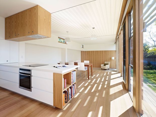 Bright open-plan kitchen and dining area with white cabinetry, a timber island and wall joinery, integrated appliances, and extensive timber-framed glazing along one side. The space opens toward a small deck and garden, with strong sunlight creating striped shadows across the timber floor. A dining table and a small seating area are visible beyond the kitchen.