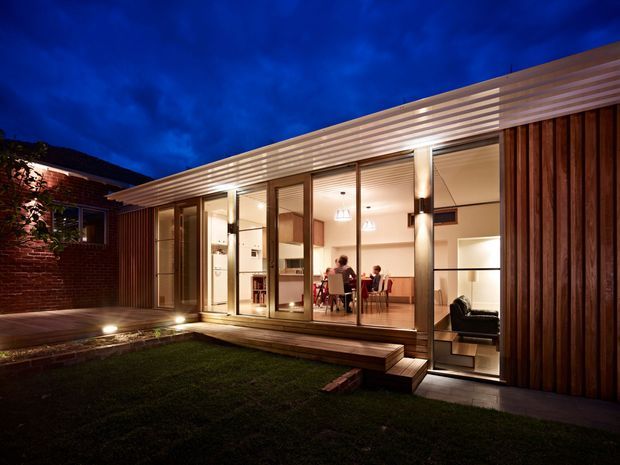Twilight exterior view of a contemporary residential extension opening onto a small landscaped courtyard. The building features extensive floor-to-ceiling glazing, warm vertical timber cladding, a projecting horizontal slatted roof canopy, timber decking and integrated exterior lighting. A dining area and living space are visible through the glass, with an adjoining brick structure at left.