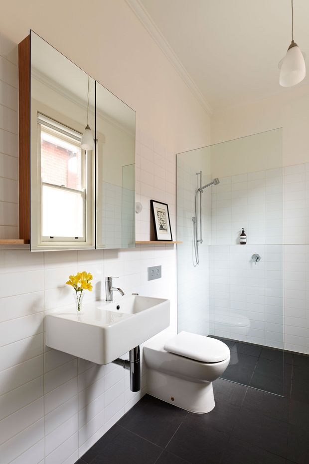 Bright, compact contemporary bathroom with white tiled walls, dark charcoal floor tiles, a wall-mounted white basin, wall-hung toilet, frameless glass shower screen, chrome fixtures, and a large mirrored cabinet. A small window and pendant lights provide natural and ambient illumination.