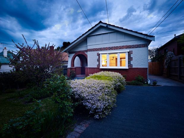 Twilight exterior photograph of a compact single-storey period home with a prominent front gable, painted white masonry, red brick detailing and a dark red brick base. The facade features a deep arched entry porch, decorative gable boarding, timber-framed windows glowing warmly from within, and a landscaped front garden beside a dark paved driveway. Overhead utility wires and neighbouring homes are visible.