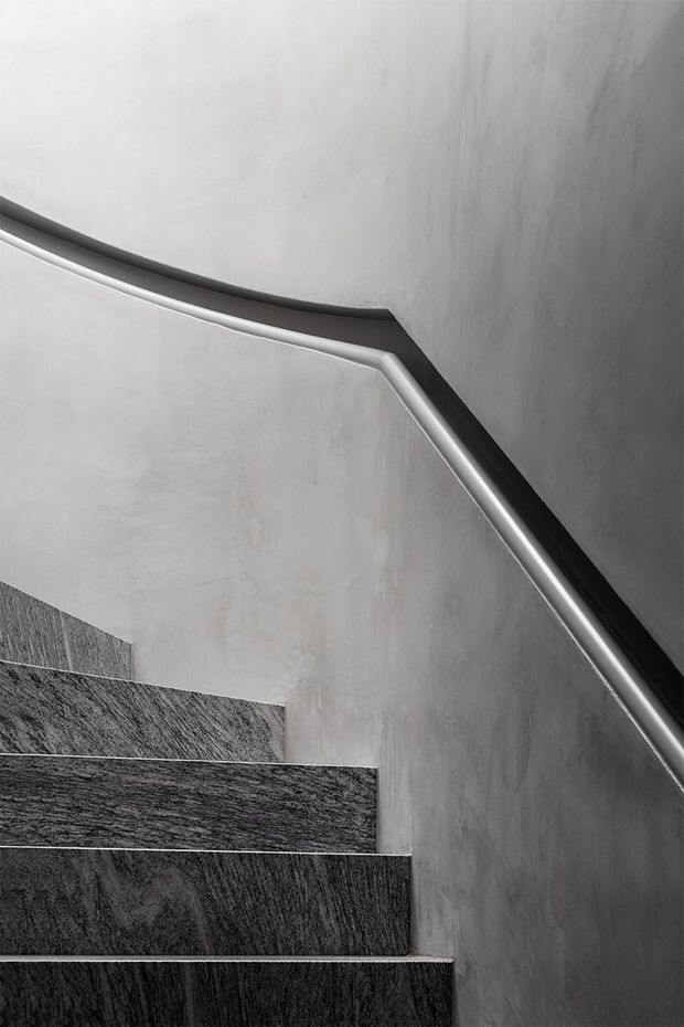 A close-up monochrome view of a contemporary stairway ascending around a corner. The stairs have dark textured stone or porcelain treads and risers, smooth light grey plastered walls, and a slim contrasting handrail with dark trim. The composition emphasizes geometric lines, material texture, and understated detailing.