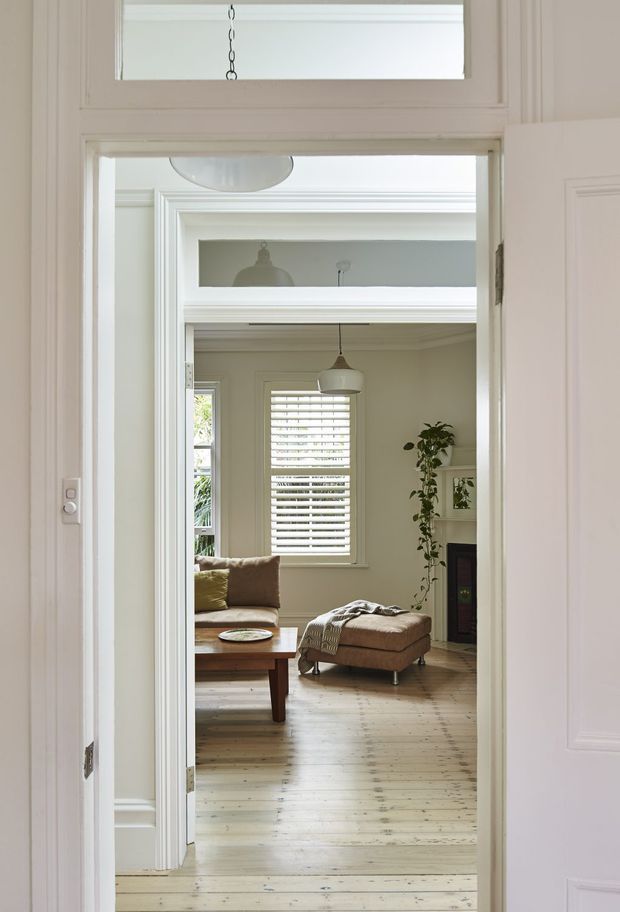 A bright view through a wide interior doorway into a refined living room. The room features pale painted walls with substantial decorative mouldings, a timber plank floor, a central pendant light, plantation shutters, a fireplace, a trailing indoor plant, and relaxed seating including a brown ottoman and sofa. The foreground doorway and transom emphasize the layered connection between rooms.