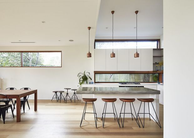 Bright open-plan kitchen and dining area with a long stone island, four bar stools, white built-in cabinetry, warm timber flooring, pendant lights, and horizontal timber-framed windows. A dining table and chairs sit to the left, with greenery visible outside.