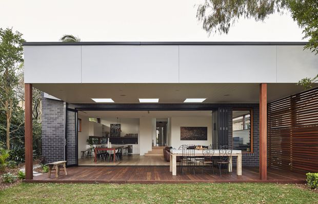 Single-storey contemporary rear extension viewed from the garden, with a broad flat roof, white rendered or panelled fascia, dark brick side walls, timber posts, and extensive openable glazing. The covered outdoor deck connects directly to an open-plan dining and living area, with a large dining table inside and a secondary outdoor dining table. Horizontal timber screening defines the right side, while roof skylights bring light into the covered space.