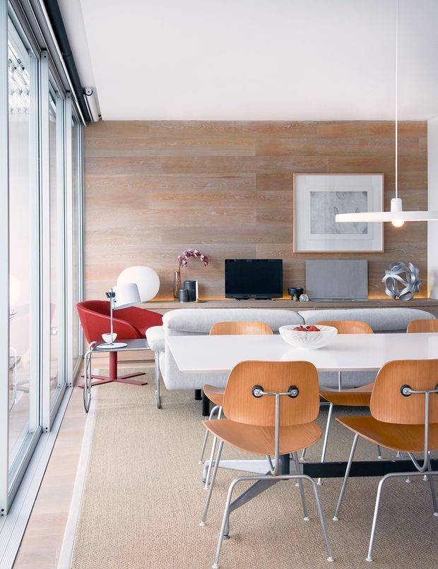 Bright, modern open-plan dining and living space with floor-to-ceiling glazing along one side, a light timber feature wall, white dining table, sculptural timber dining chairs, and a grey sofa. Mid-century-inspired furniture, minimalist artwork, and a linear pendant light create a refined contemporary interior.
