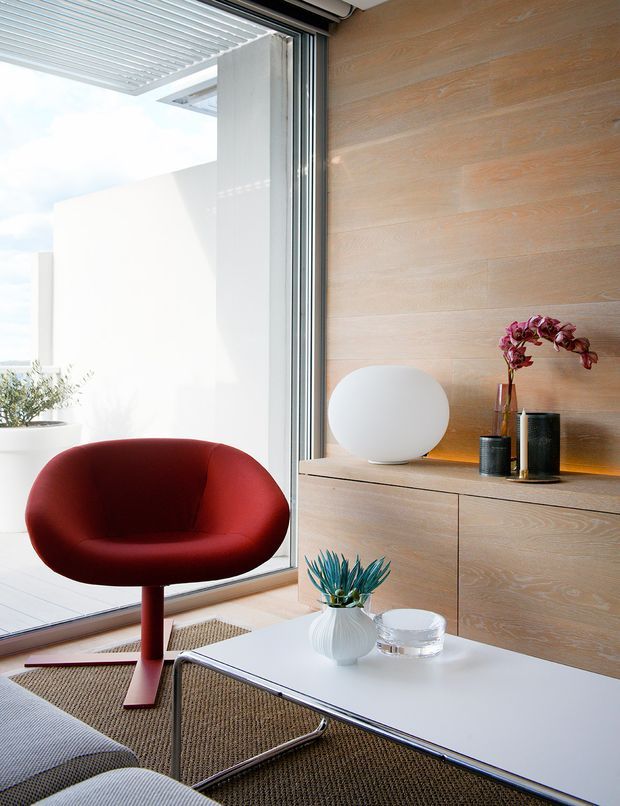A bright, refined modern living room corner with a sculptural deep-red lounge chair, white coffee table, and low timber built-in cabinetry against a horizontally panelled timber wall. Floor-to-ceiling sliding glazing opens toward a light outdoor terrace, with a slatted overhead screen visible outside. Minimal decorative objects and indoor plants add subtle accents.