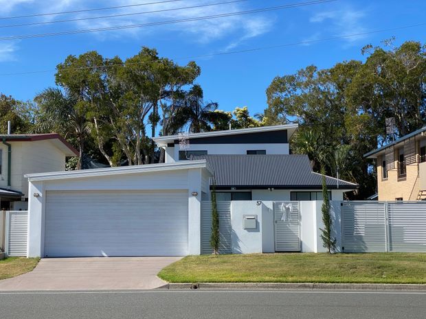 Street-facing view of a contemporary two-storey residence with a white rendered facade, broad double garage, dark grey corrugated metal skillion-style roof, horizontal slatted fencing, and a small lawn. The upper level has dark cladding and narrow horizontal windows, while mature trees and neighbouring homes frame the property.