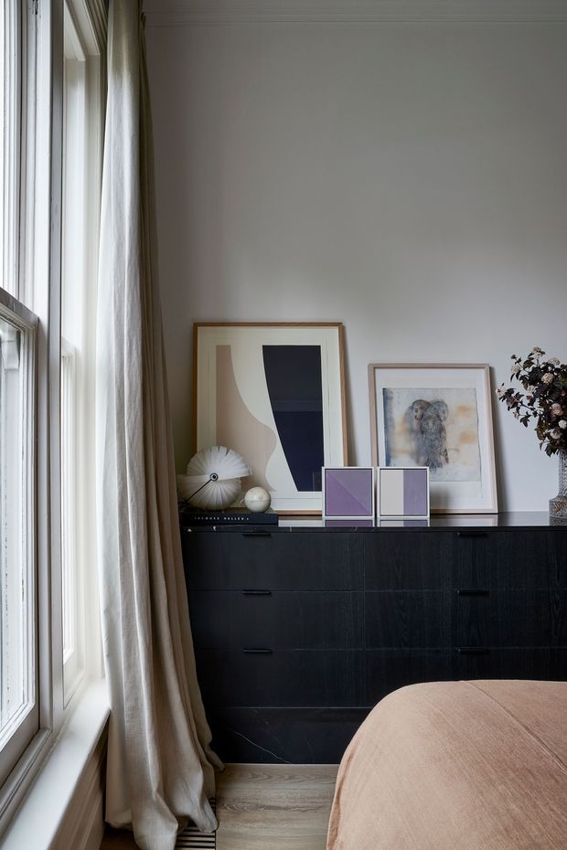 A softly lit, refined bedroom corner with tall white-framed windows and full-length neutral linen curtains. A black stained timber sideboard with minimalist hardware sits against a pale wall, styled with framed abstract artwork, sculptural decor and dried foliage. The edge of a rounded upholstered bed is visible in the foreground, with light timber flooring and a calm neutral palette.