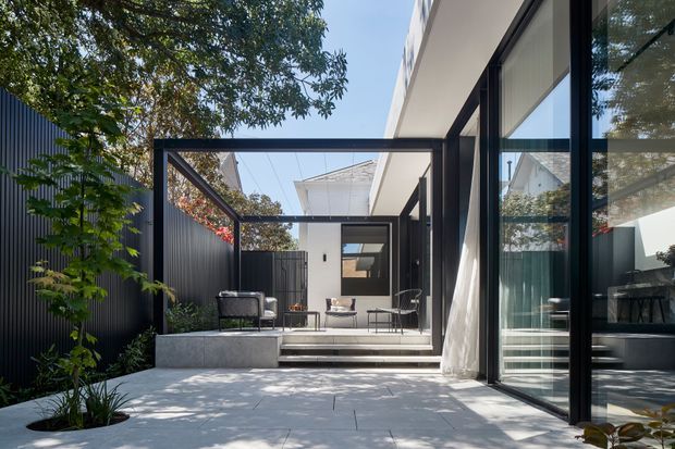 A contemporary residential courtyard or terrace viewed from outdoors, with pale paved flooring, a raised seating platform, black-framed floor-to-ceiling glazing, and a slim black pergola-like structure. The space includes minimal outdoor lounge furniture, lush planting along a dark privacy fence, and strong indoor-outdoor visual connection through open glass doors. Bright daylight and clean linear detailing create a refined, airy atmosphere.