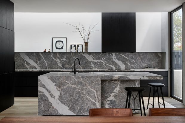 A sophisticated contemporary kitchen with a large grey veined stone island and matching full-height splashback. Matte black cabinetry runs along the rear wall and beside the island, contrasting with white upper walls and light timber flooring. Two black bar stools sit at the island, while a dining table occupies the foreground. Full-height glazing at the side brings in natural light and views to an exterior fence and vegetation.