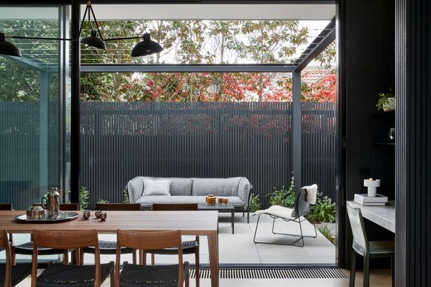 A contemporary dining and living area opening onto a private paved courtyard through expansive floor-to-ceiling glazing. The interior features a timber dining table and chairs, a grey sofa, a sculptural lounge chair, black pendant lighting, and dark vertical wall panelling with open shelving. The courtyard has a dark slatted privacy fence, greenery, and a covered glazed structure, creating a strong indoor-outdoor connection.