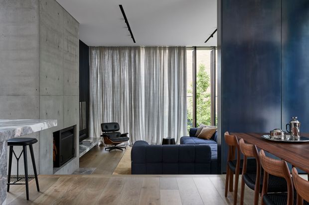 A contemporary open-plan living and dining area with a dark blue built-in wall and timber dining table in the foreground. The living zone features a deep blue sofa, black leather lounge chair, sheer full-height curtains, a concrete fireplace wall, timber flooring, and large glazed openings overlooking greenery. Linear black ceiling track lights and restrained furnishings create a refined, moody interior.