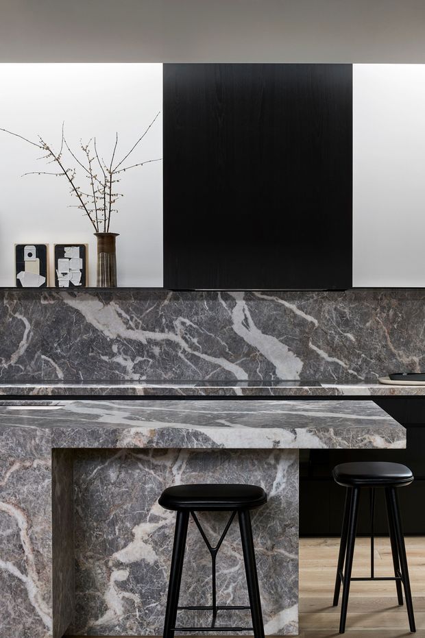 A refined contemporary kitchen centered on a dramatic grey and white veined stone island and matching backsplash. Matte black cabinetry and a large black overhead panel create a strong graphic contrast against pale walls and light timber flooring. Two black stools sit at the island, while a simple branch arrangement and framed objects add restrained decoration.