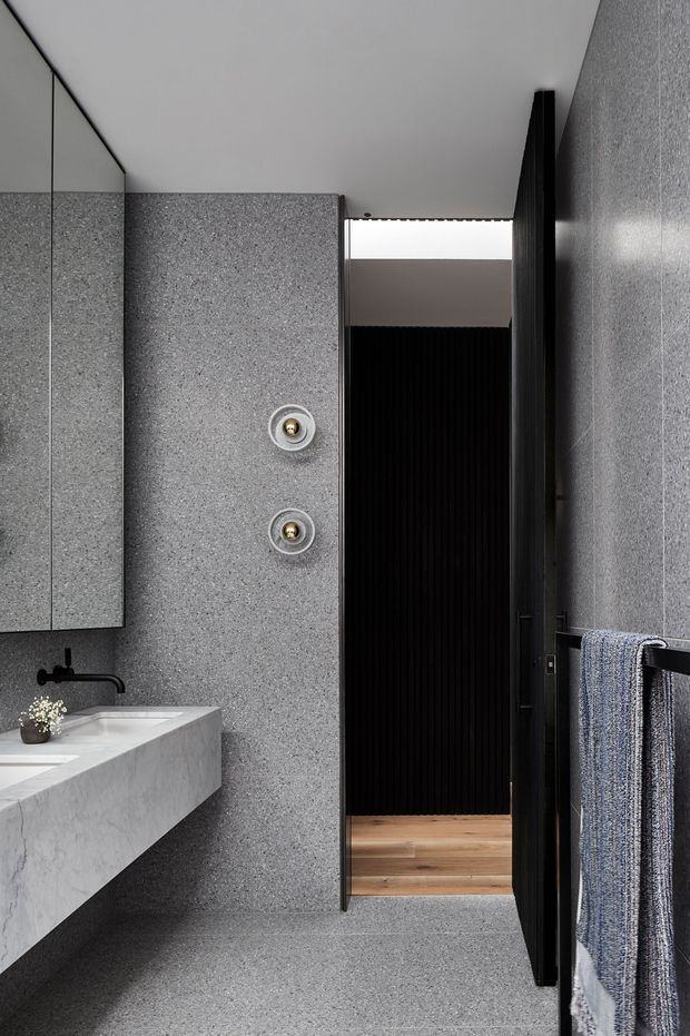 A refined contemporary bathroom with grey stone-look wall and floor surfaces, a floating pale stone vanity, black wall-mounted tapware, round brass-accented controls, a large mirrored cabinet, and a dark ribbed doorway leading to a timber-floored adjoining area. A towel hangs on a black rail at right.
