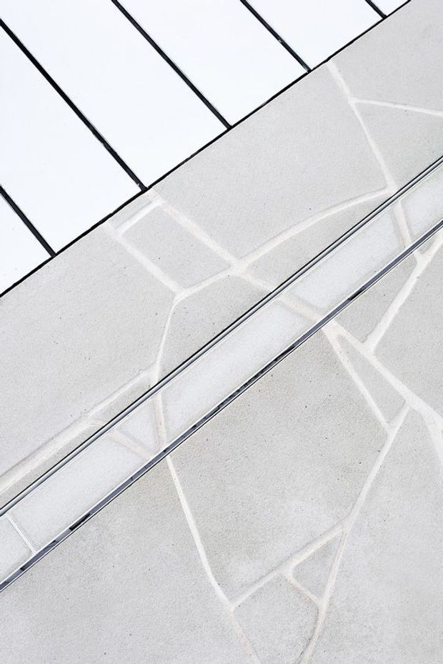 Abstract close-up of a pale grey tiled architectural surface with dark grout lines and a long narrow linear metal drain or channel crossing diagonally. Bright reflected geometric lines create a clean, minimal composition.