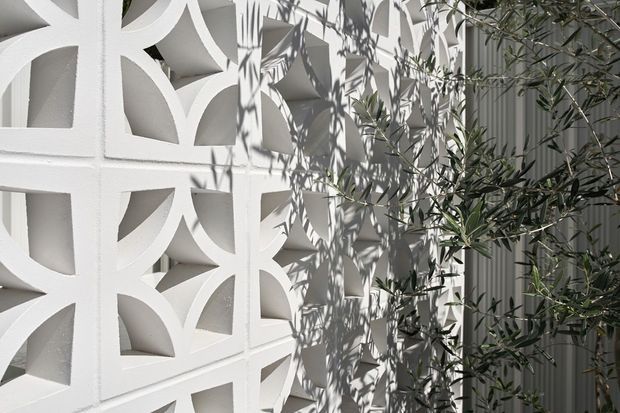 Close-up exterior architectural detail featuring a white decorative breeze-block screen with repeating curved geometric openings. Strong sunlight creates layered shadows from the screen and nearby olive-like foliage across a pale wall, with vertical ribbed cladding visible to the right. The image emphasizes facade texture, pattern, and filtered light.